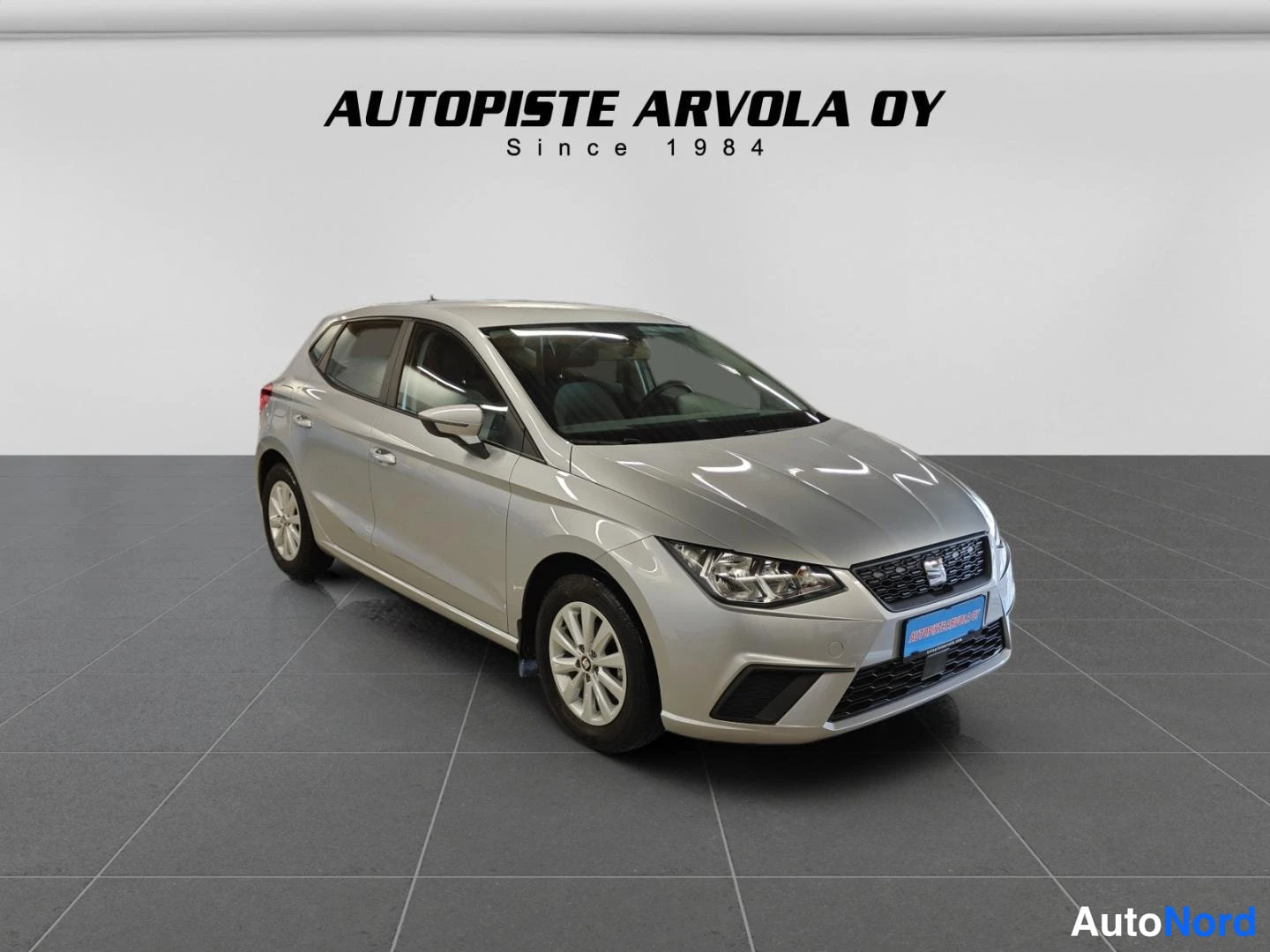 Etuviisto, nokka oikealle: Seat Ibiza -henkilöauto hopeanvärisenä kuvattuna myymälätilassa, kolmen neljännestä keula oikealla.