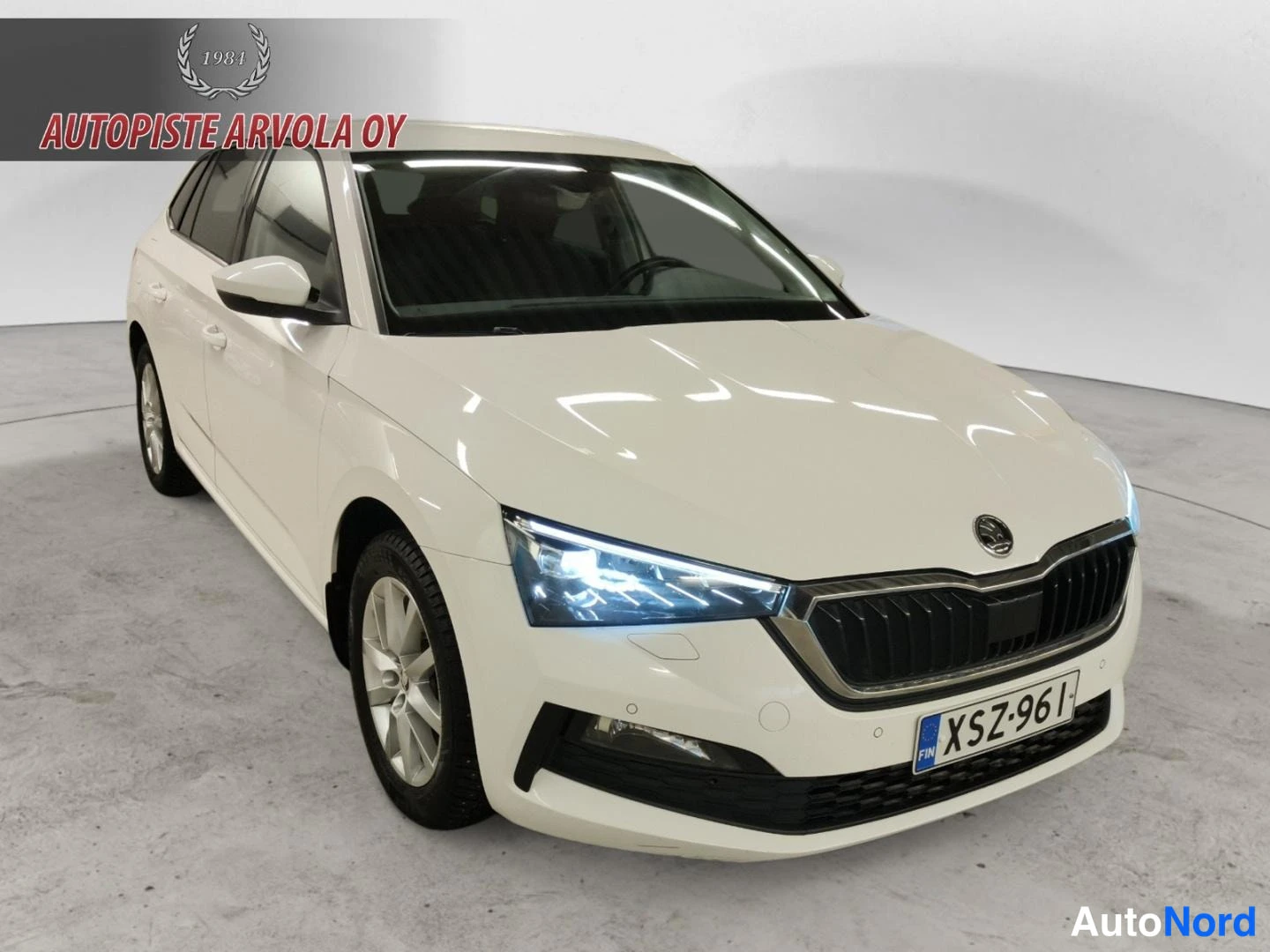 Etuviisto, nokka oikealle: Valkoinen Skoda Scala farmari etuviistöstä oikealta, LED-ajovalot päällä, rekisteritunnus.