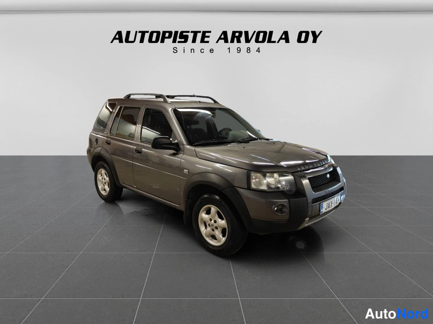 Etuviisto: Land Rover Freelander tummanharmaana etukolmesta-neljänneskuvassa studiossa; katolla kattokaiteet, rekisterikilpi JA X-1 0 näkyvät, taustalla Autopiste Arvola O
