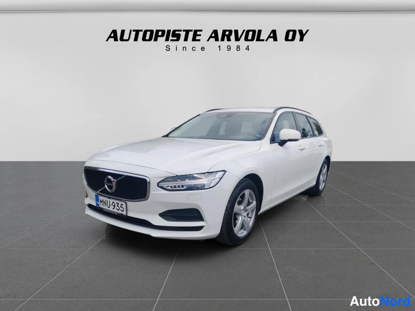 Etuviisto, nokka vasemmalle: Valkoinen Volvo V90 -farmariauto kulmassa etuosa vasemmalle päin valkoisella studiotakeella, rekisterikilpi, taustalla AUTOLIPASTE ARVOLA OY -teksti.
