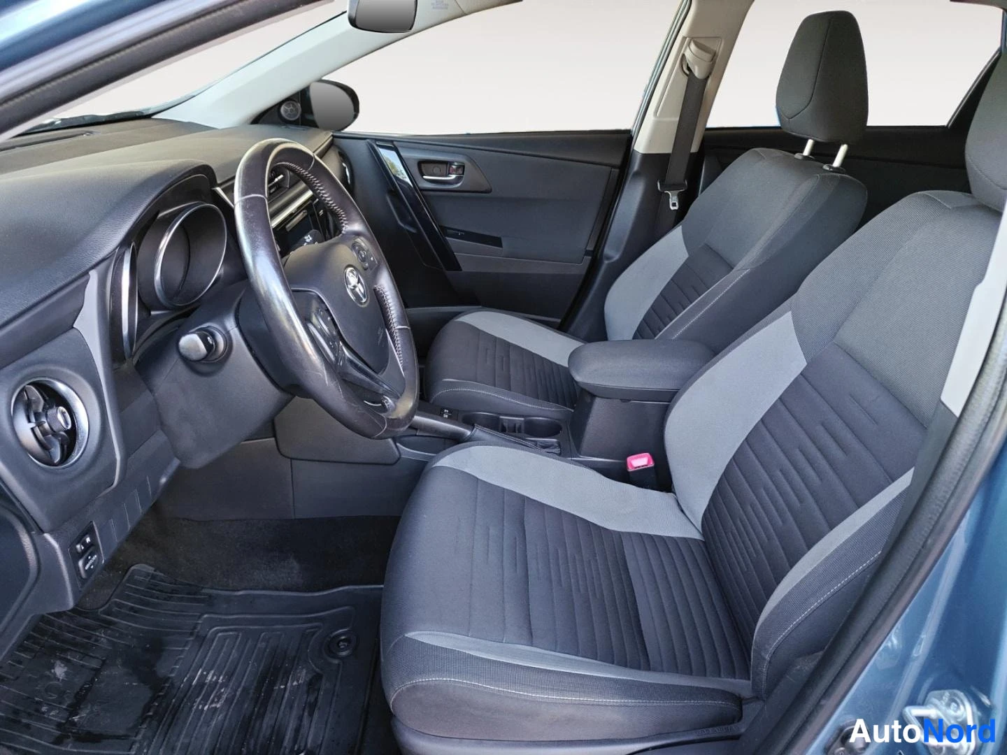Etuviisto: Auton ohjaamo ja etupenkku: tumma sisusta, Toyota-merkkiohjauspyörä, tummat kangasistuimet harmailla sisustoilla.