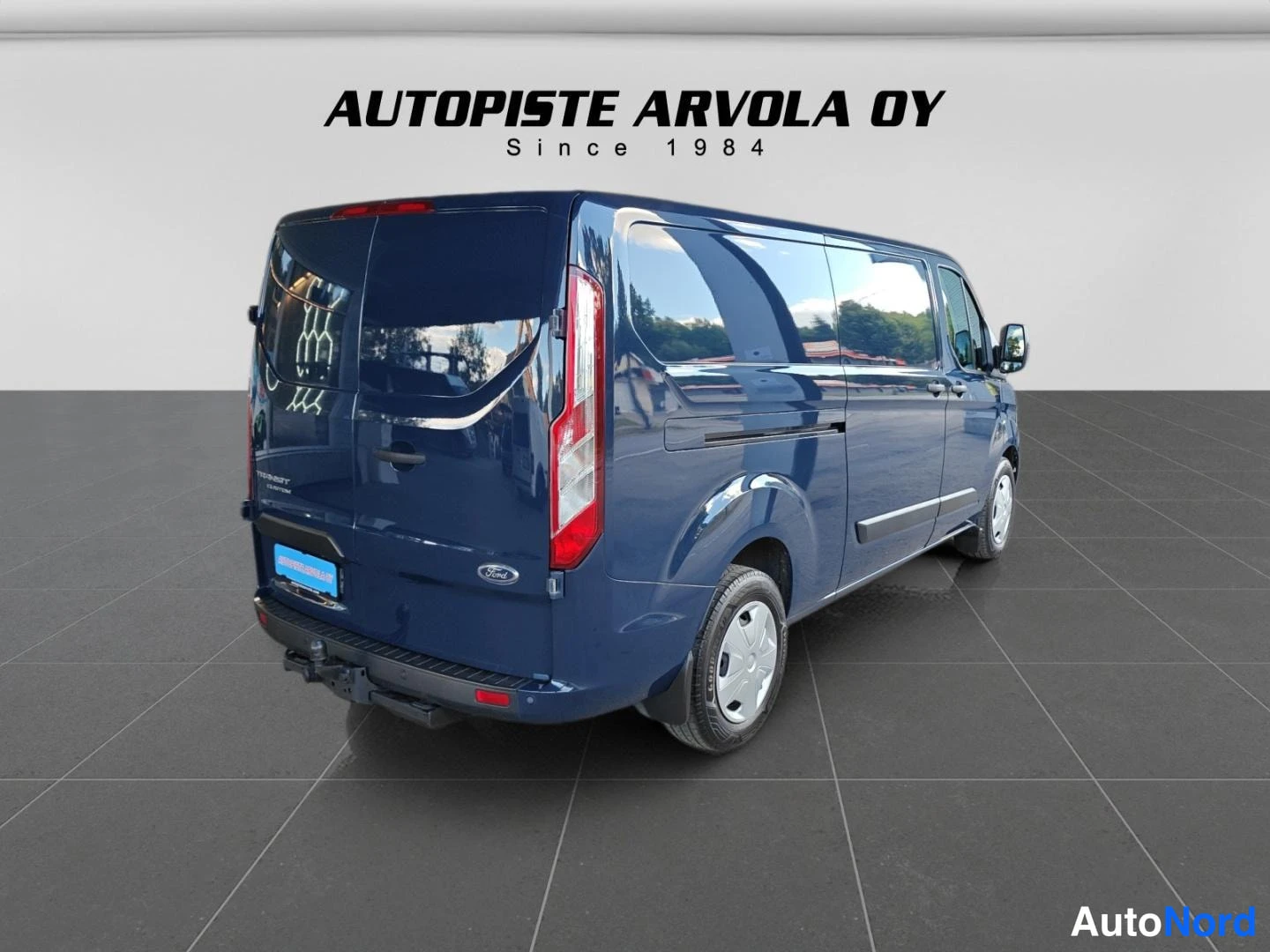 Takaviisto: Sininen pakettiauto takavartinurkkakuvassa sisätiloissa, Autopiste Arvola Oy -tausta.
