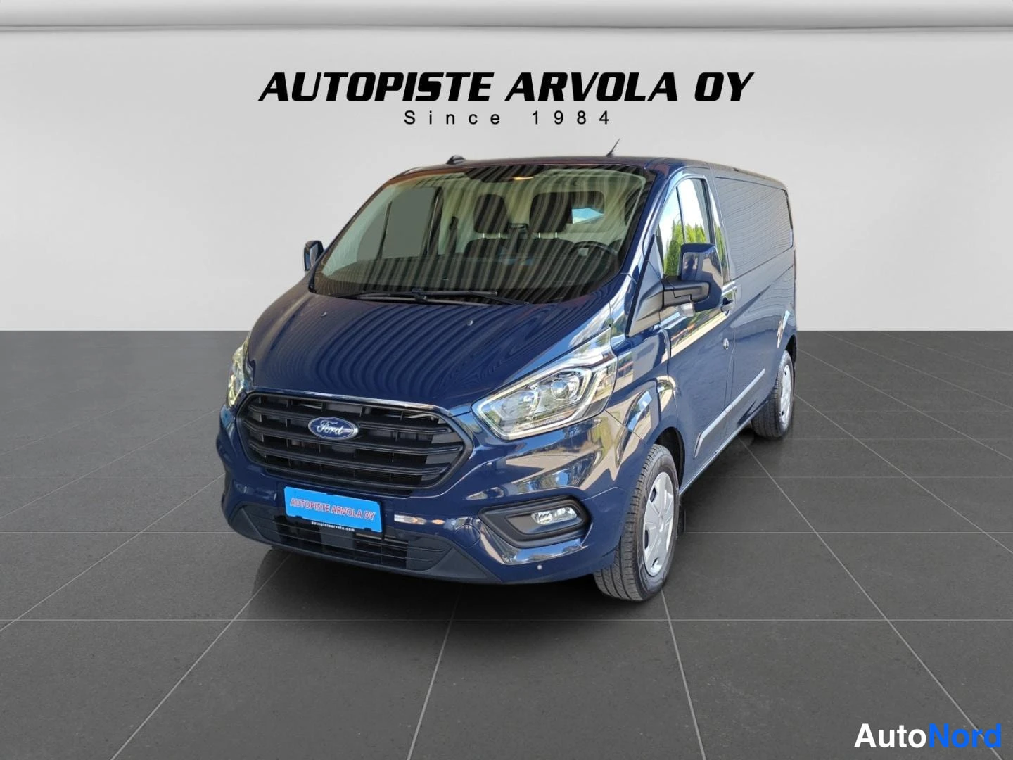 Etuviisto, nokka vasemmalle: Sininen Ford Transit Custom -pakettiauto edestä vasemmalta, studio-tyylinen kuva Autopiste Arvola Oy -taustalla.