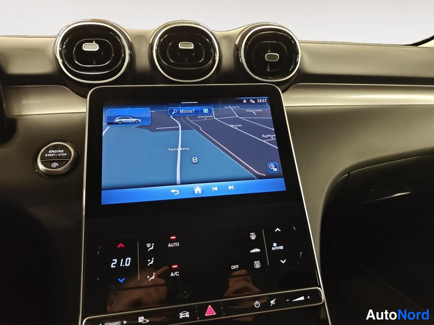Edestä: Infotainment-näyttö näyttää navigointikarttaa hakukentällä 'Minne?'; alla ilmastointipaneeli asetuksineen ja ENGINE START/STOP -painike vasemmalla.