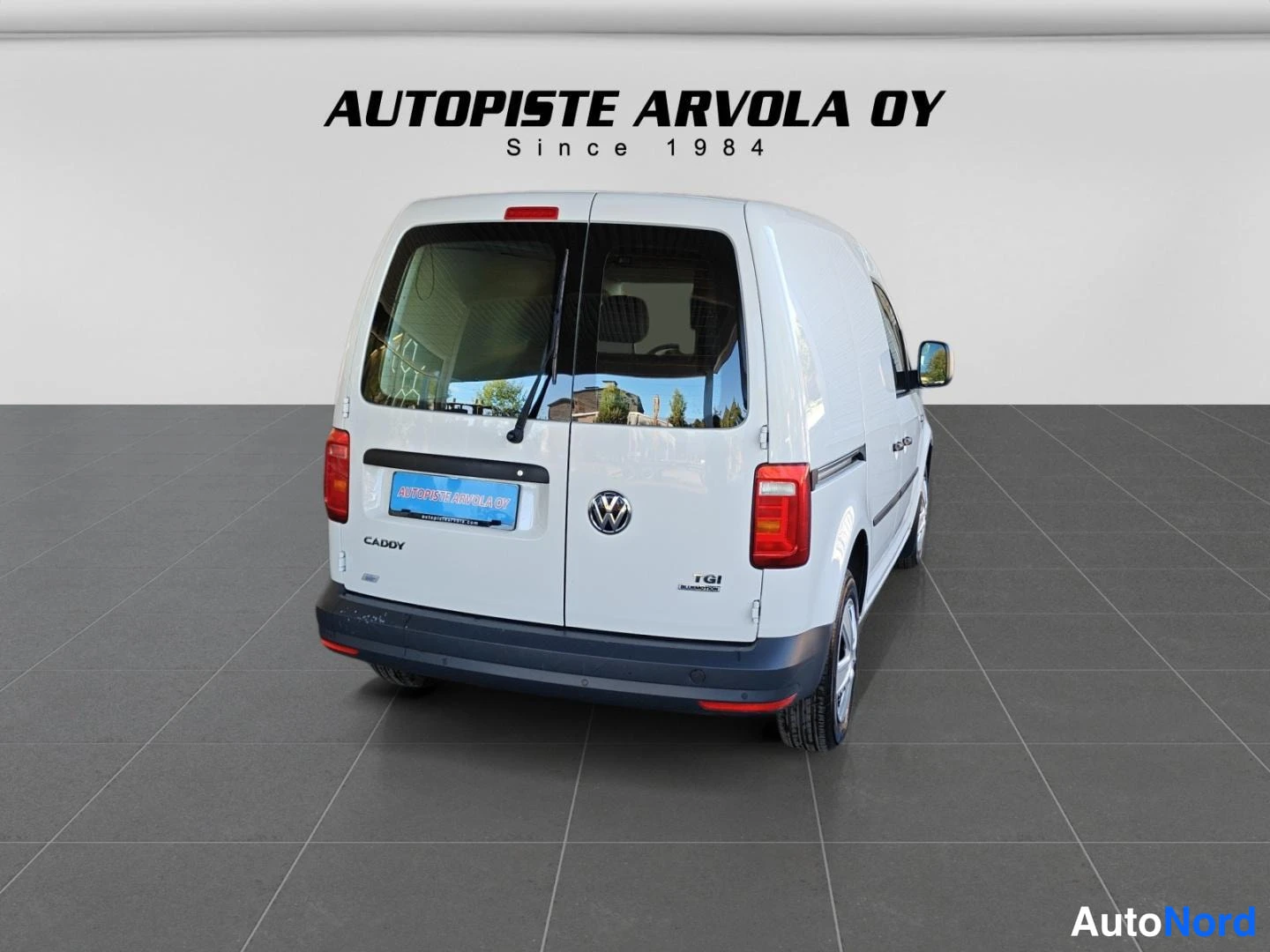 Takaviisto: Valkoinen Volkswagen Caddy -pakettiauto takaa kuvattuna, takaluukussa VW-merkki ja TGI Bluemotion-teksti, sininen Autopiste Arvola -kilpi puskurissa.