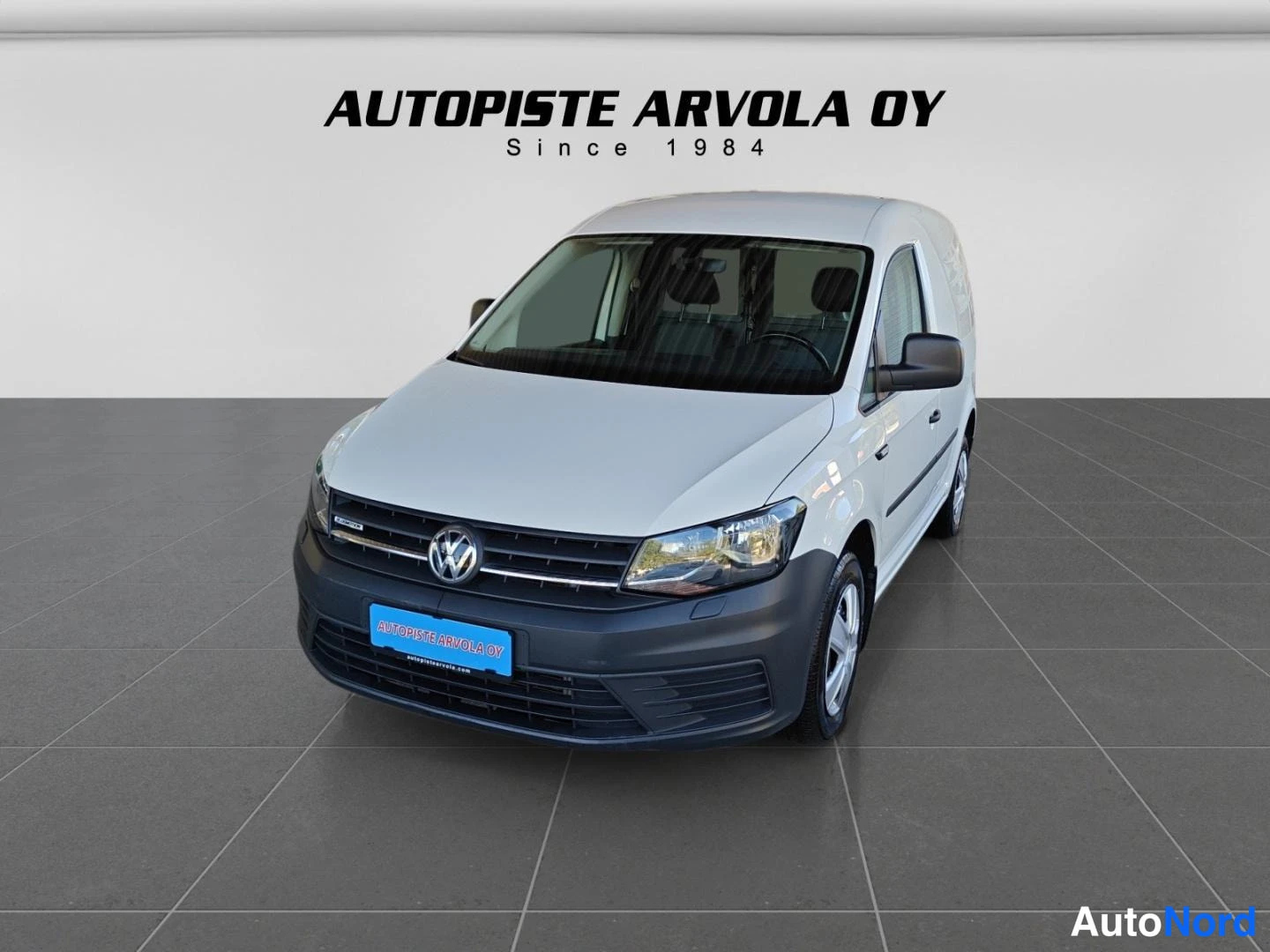 Etuviisto, nokka oikealle: Valkoinen Volkswagen Caddy -pakettiauto etuviistosta kuvattuna, tumma etupuskuri ja sininen Autopiste Arvola -kilpi grillin alla.