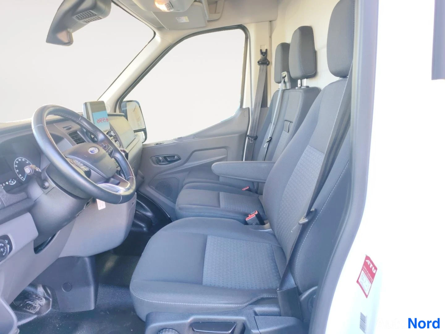Edestä: Paku Ford Transitin ohjaamosta sisältä otettu kuva: ohjauspyörä, kojelauta ja tummat kankaanistuimet; kuvan oikea reuna on kirkkaasti ylipaljastunut.