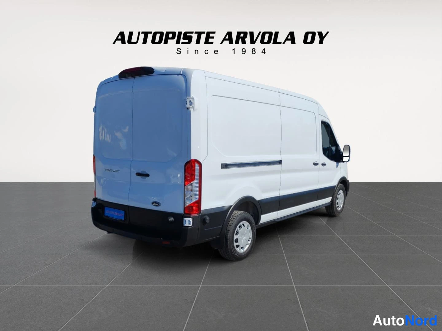 Takaviisto: Valkoinen Ford Transit -pakettiauto takaviistosta valkoista seinää vasten: takana kaksoisovi, kyljessä liukuovi; kuvassa mainos tekstillä 'AUTOPISTE ARVOLA OY S