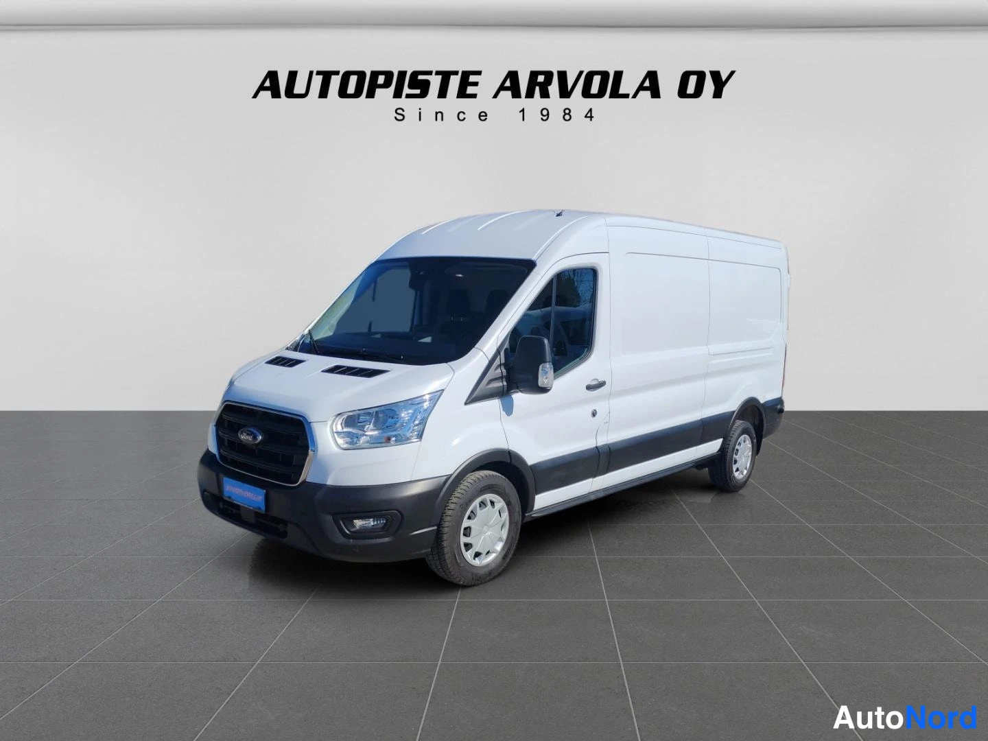 Etuviisto, nokka vasemmalle: Valkoinen Ford Transit -pakettiauto etuviistosta studio­ympäristössä: valkoinen kori, tummat puskurit, kyljessä liukuovi; taustalla teksti 'AUTOPISTE A