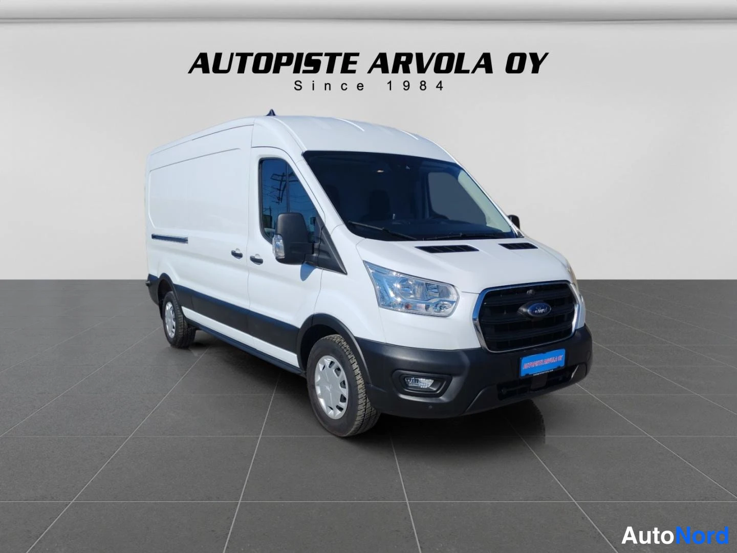 Etuviisto, nokka oikealle: Valkoinen Ford Transit -pakettiauto kuvattu kolmannenvälinen etunäkymä, keula oikealla, Autopiste Arvola Oy -studiovaalea tausta.