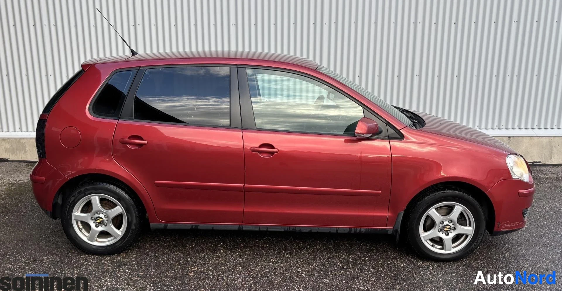 Sivulta: Vasemman sivun näkymä punaisesta viisiporttisesta hatchbackista. Autossa on hopeiset kevytmetallivanteet ja takakaton antenni. Auto seisoo harmaalla asfaltilla