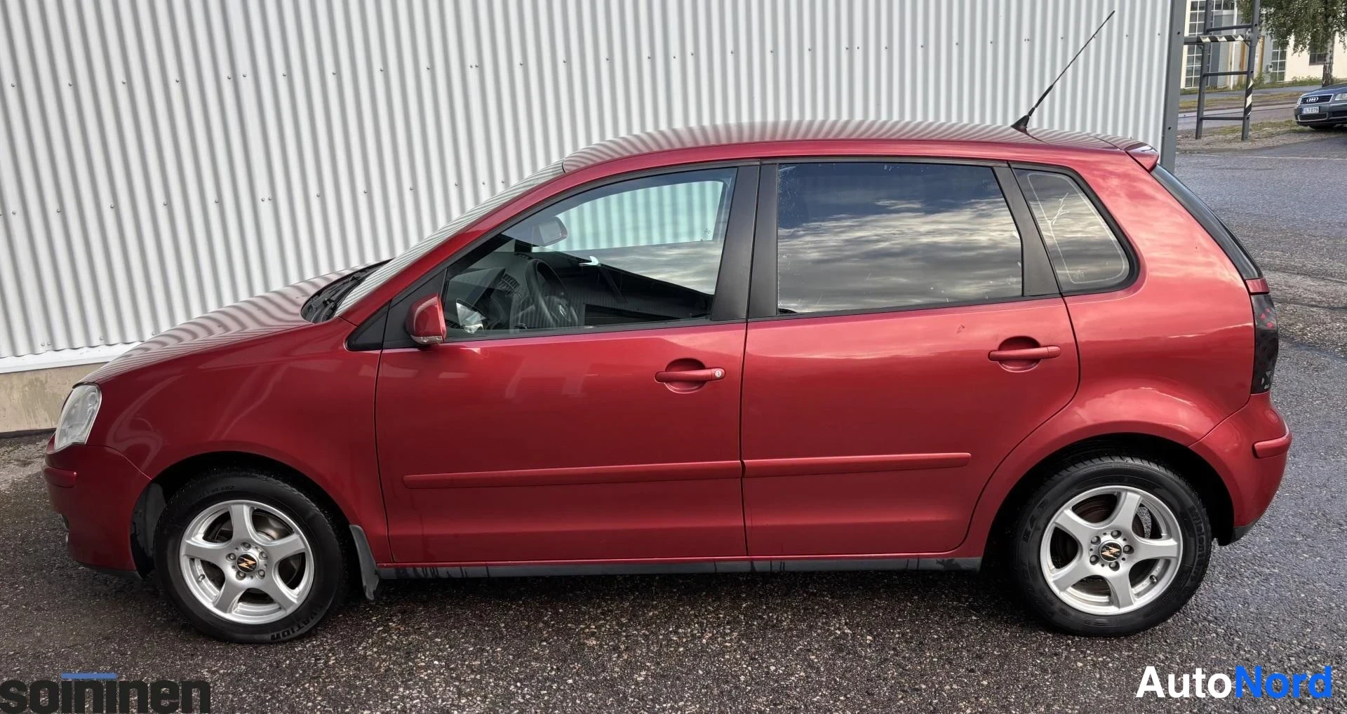 Sivulta: Oikean sivun näkymä punaisesta viisiporttisesta hatchbackista. Autossa on hopeiset kevytmetallivanteet ja takakaton antenni. Auto seisoo harmaalla asfaltilla ko