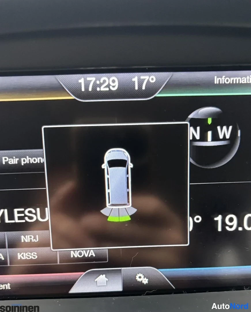 Yksityiskohta: Ford SYNC-näytön lähikuva: auton kuvasymboli pysäköintiantureilla, kello 17:29, lämpötila 17 astetta ja YLESUOMI-radiolähettien valintapaneeli.