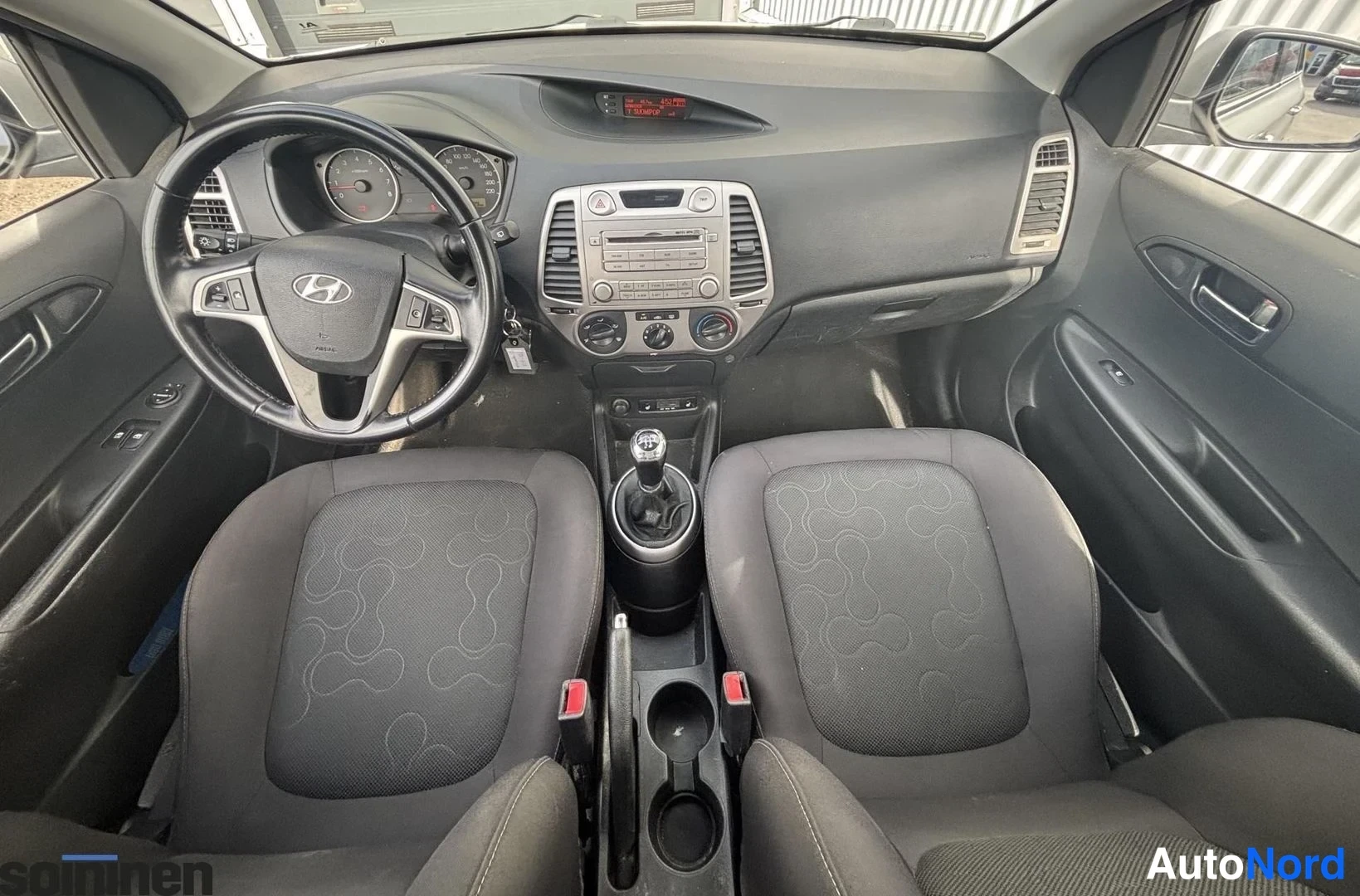 Takaviisto: Hyundai i20:n kojelauta ja ohjaamo ylhäältä kuvattuna; ohjauspyörä, radionäyttö, ilmastointisäätimet ja manuaalivaihteiston vipu näkyvät, tummat etuistuimet auk