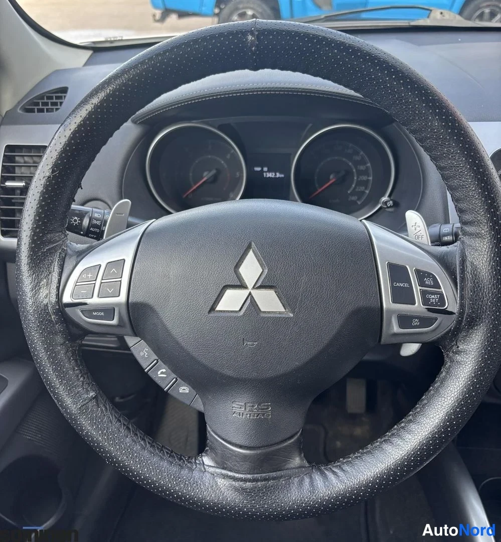 Yksityiskohta: Ohjauspyörän lähikuva Mitsubishi-logo keskellä, varustettuna monitoiminapeilla. Mittareiden tausta näkyy.
