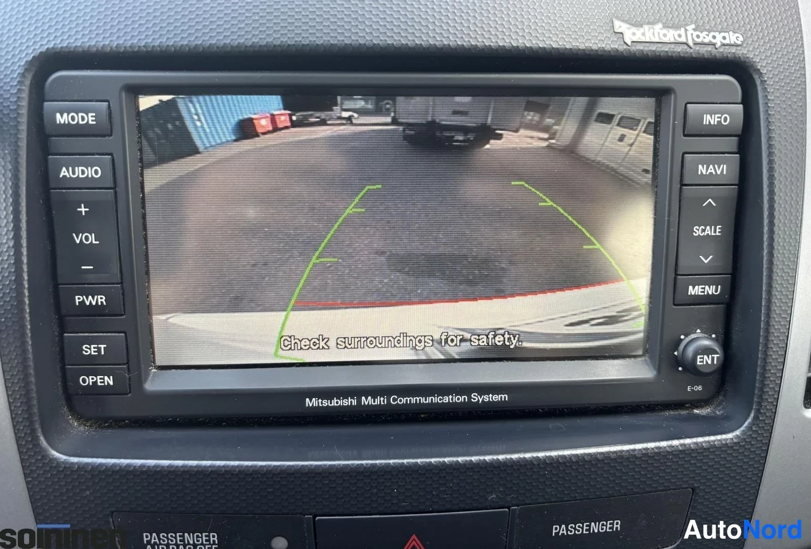 Muu: Mitsubishi Multi Communication System -näyttö peruutuskameran kuvalla ja tekstillä 'Check surroundings for safety.'