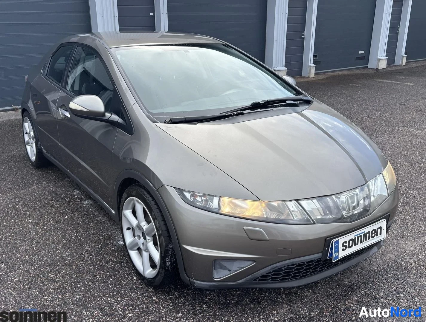 Etuviisto, nokka oikealle: Harmaankirjava Honda Civic viistoperä kuvattu kulmasta keula oikealla, hopeavanteet ja soininen-tunnus etupuskurissa.