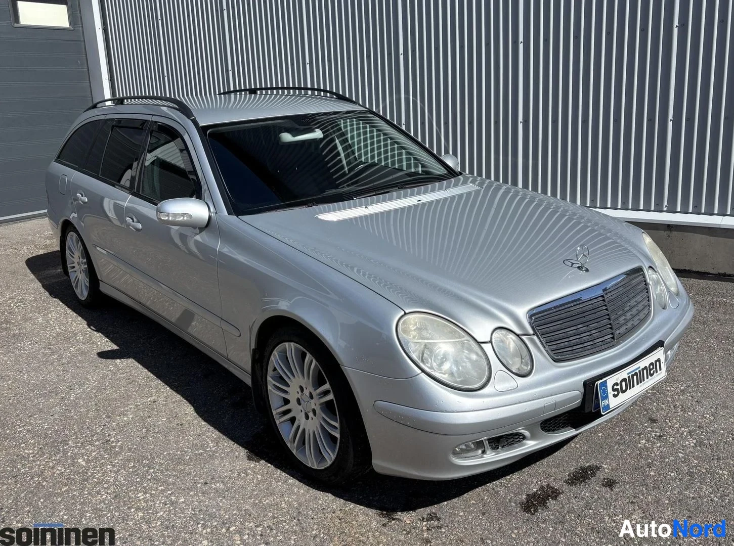 Etuviisto, nokka oikealle: Hopeanharmaan Mercedes-Benz farmarin etukulma, konepelti, etusäleikkö ja ajovalot näkyvät katuajossa teollisen hallin seinän edessä.