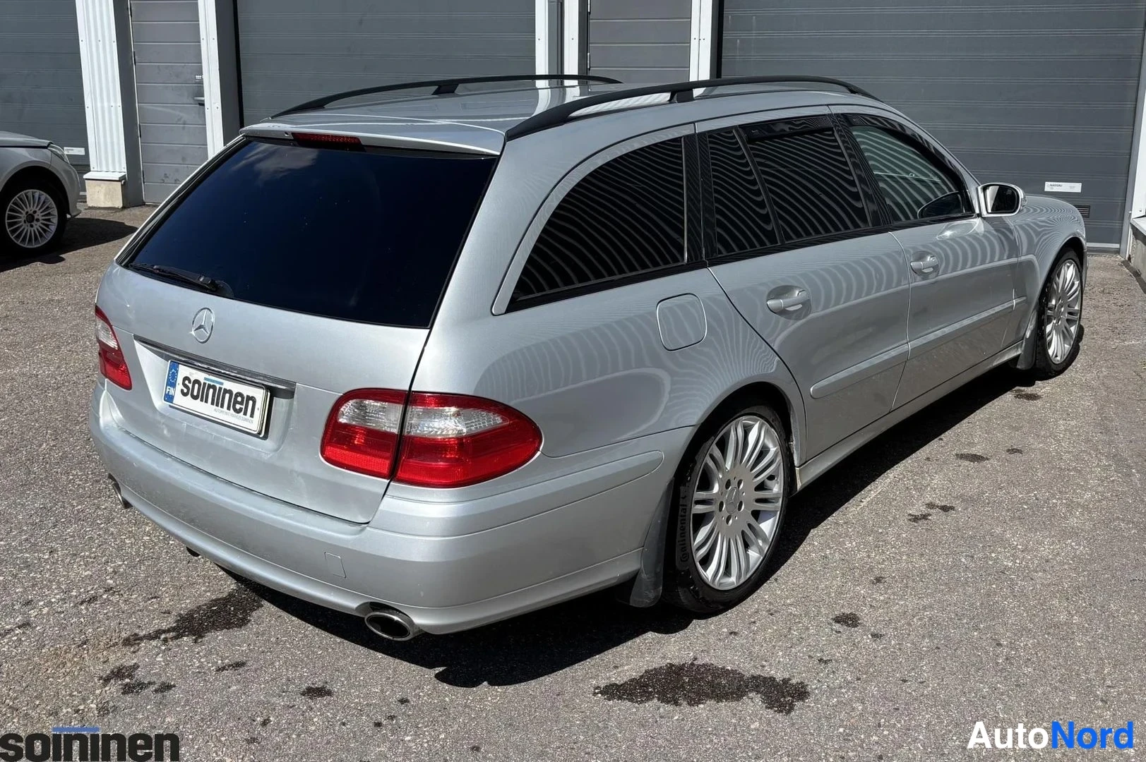 Takaviisto: Hopeanharmaan Mercedes-Benz farmarin takakulma, punaiset takavalot, pakoputki ja monivannevanteet näkyvät parkkihallissa.