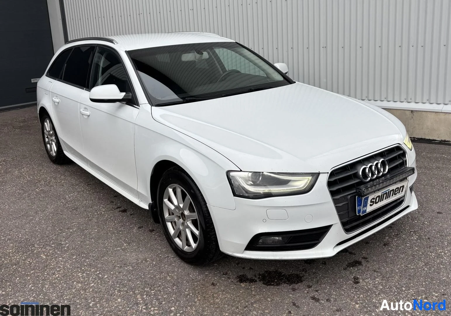 Etuviisto, nokka oikealle: Etinen ja vasen kylki näkyvissä oleva valkoinen Audi A4 Avant. Auto seisoo asfaltilla harmaan seinän edessä. Valot on päällä, metallivanteet ja katolip