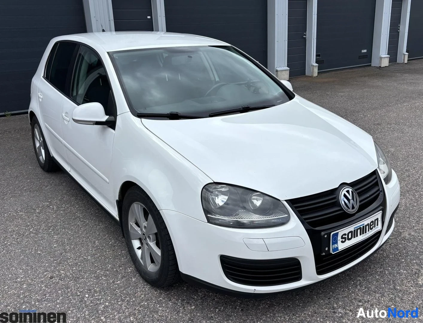 Etuviisto, nokka oikealle: Valkoinen auto oikeasta kulmasta. Etupää, jossa VW-logo ja 'soininen' rekisterikilpi.