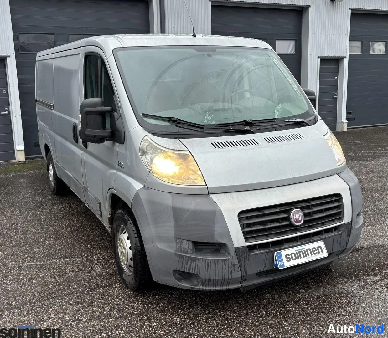 Etuviisto, nokka oikealle: Silver Fiat Ducato van, front and driver-side angle. The headlights are on (yellow). Black plastic bumper covers the lower front. A license plate with