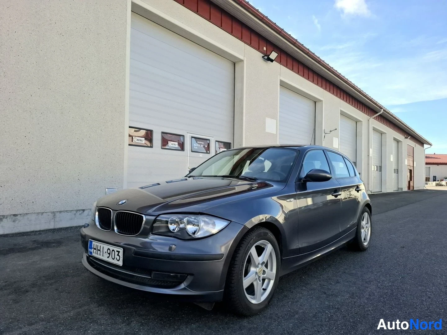 Etuviisto, nokka vasemmalle: Harmaa BMW 116 5-ovinen hatchback etuviistossa autotallin edessä, rekisteritunnus HH1-903.