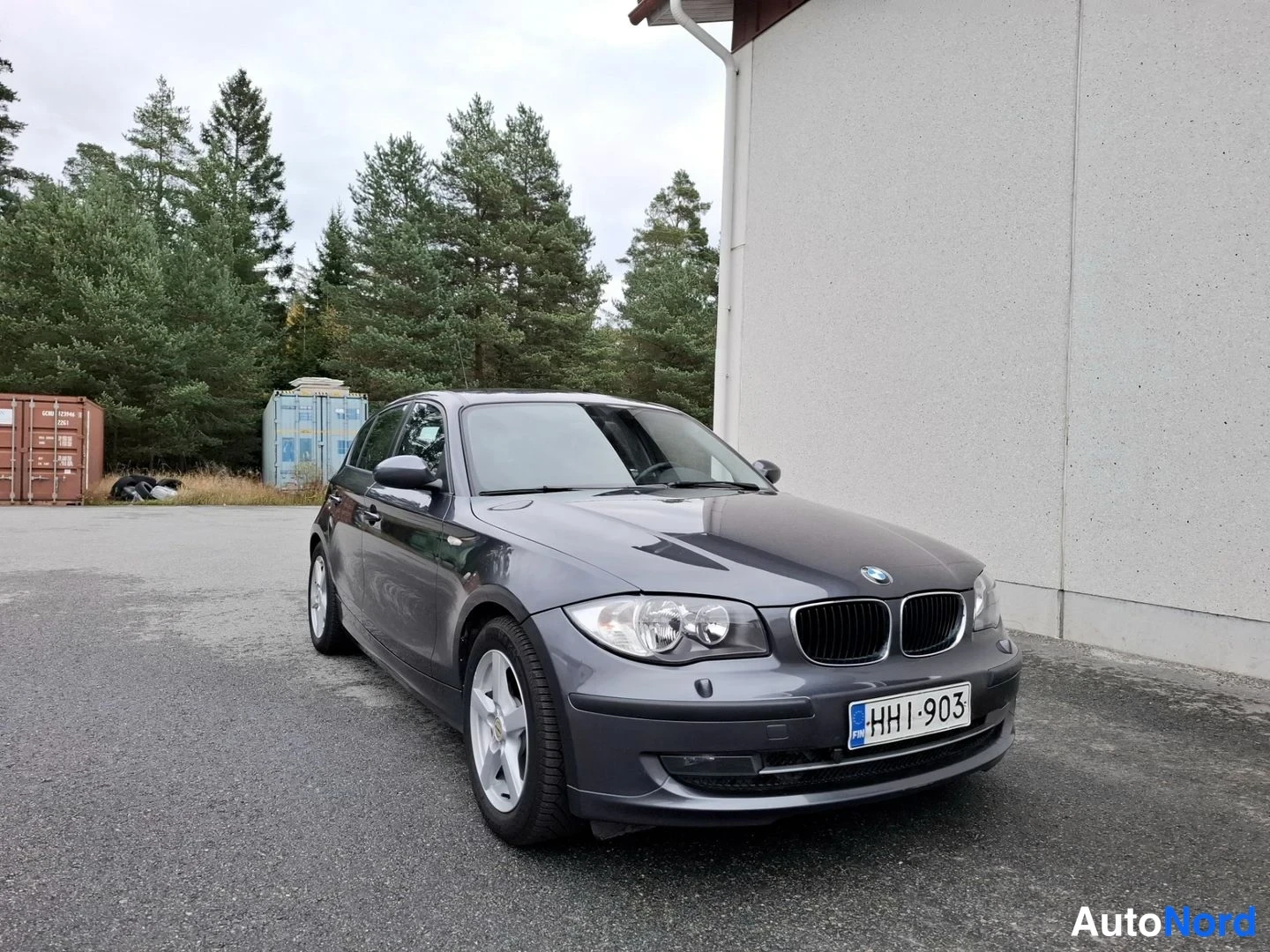 Etuviisto, nokka oikealle: BMW 1-sarjan viistoperä tummanharmaana edestä oikealta kuvattuna valkoisen rakennuksen edessä.