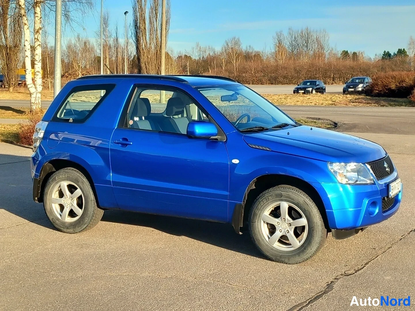 Etuviisto, nokka oikealle: Sininen Suzuki Grand Vitara kolmiovinen sivulta kolmasosaedestä, keula oikealla, kuva ajopaikassa pihamaalla.