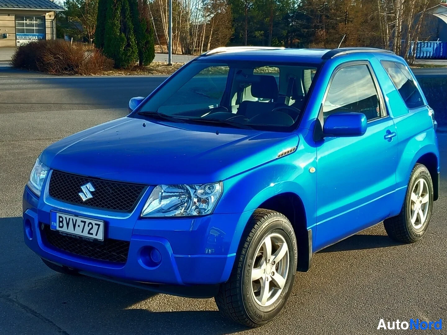 Etuviisto, nokka vasemmalle: Sininen Suzuki Grand Vitara -maastoauto kolmasosaedestä, keula vasemmalla, rekisterikilpi luettavissa.