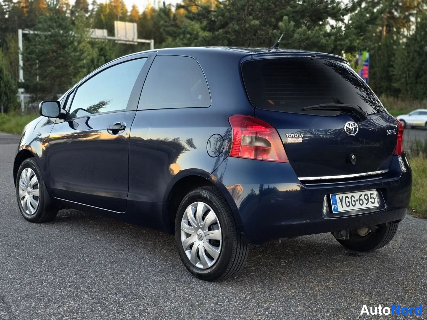 Takaviisto: Tummansininen Toyota Yaris takakolmanneltapuolelta; takaluukussa TOYOTA- ja YARIS-tekstit, Toyota-embleemi ja rekisterikilpi.