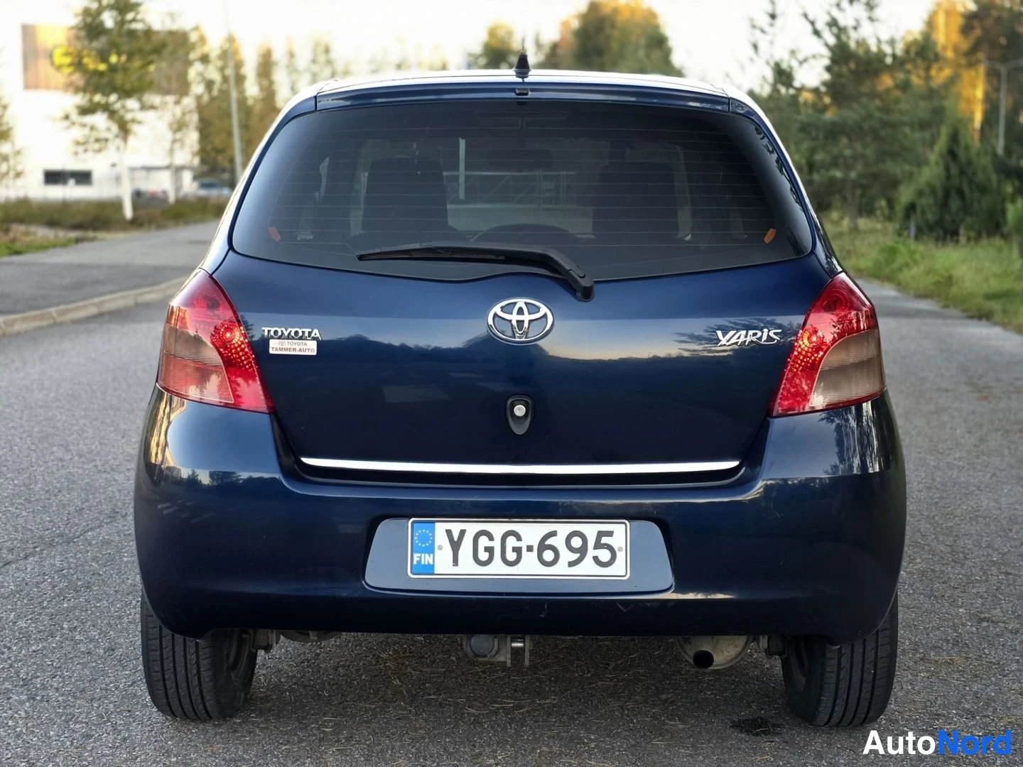 Takaa: Tummansinisen Toyota Yarisin suora takaosa; takaluukussa TOYOTA- ja YARIS-tekstit, Toyota-logo ja rekisterikilpi, takalasinpyyhkijä näkyvissä.