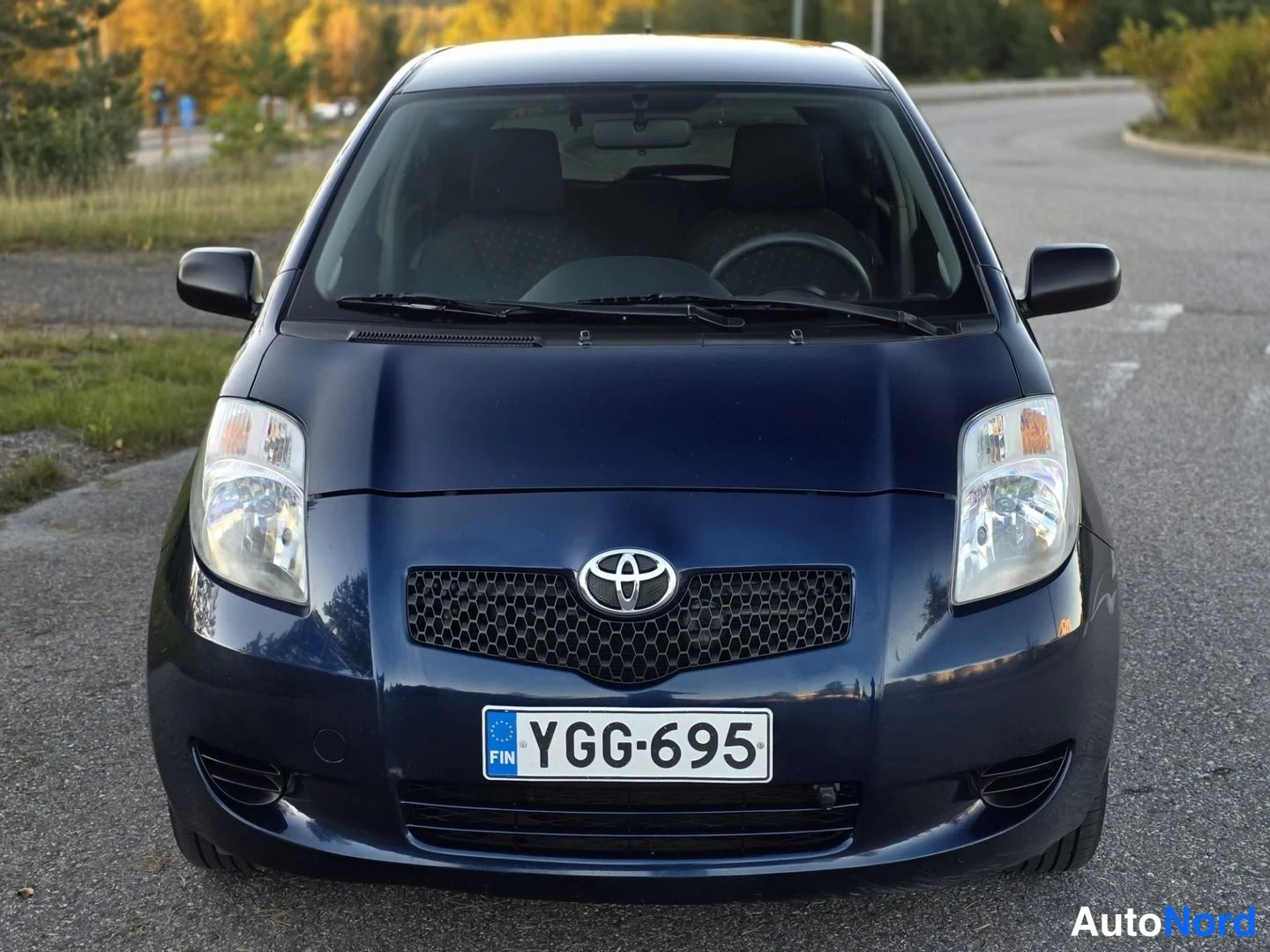 Edestä: Toyota Yarisin suora etunäyttö: tummansininen auto, musta verkkosäleikkö Toyota-merkillä, molemmat ajovalot ja -rekisterikilpi näkyvät.