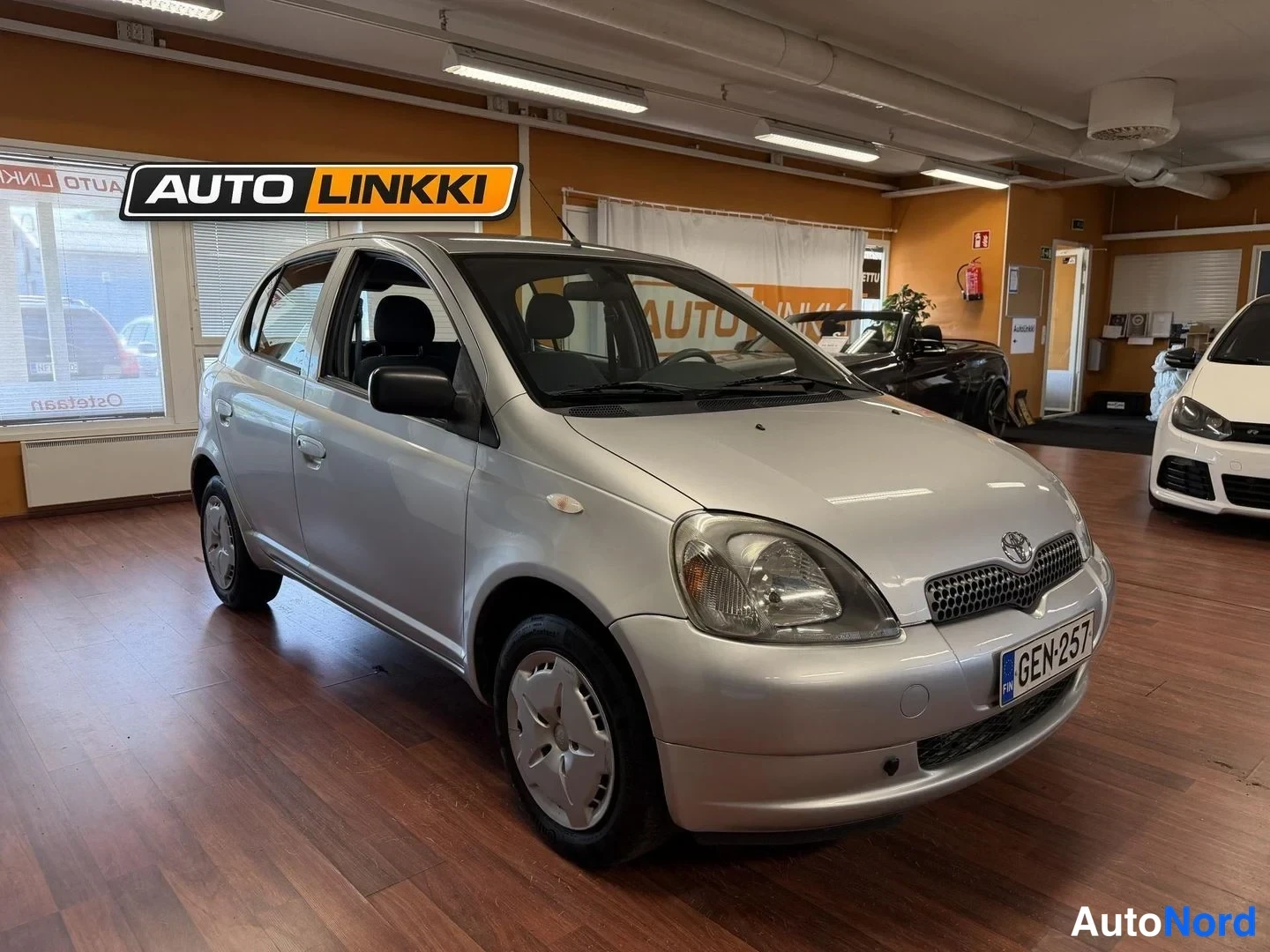 Etuviisto, nokka oikealle: Hopea Toyota Yaris -henkilöauto nähty kolmannelta etukulmalta, keula oikealla, Autoluokan myyntitilassa.