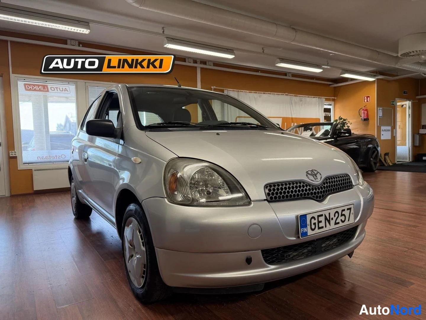 Etuviisto, nokka oikealle: Hopea Toyota Yaris -henkilöauto etukulmasta, keula oikealla, rekisterikilpi, myyntitilan sisällä.