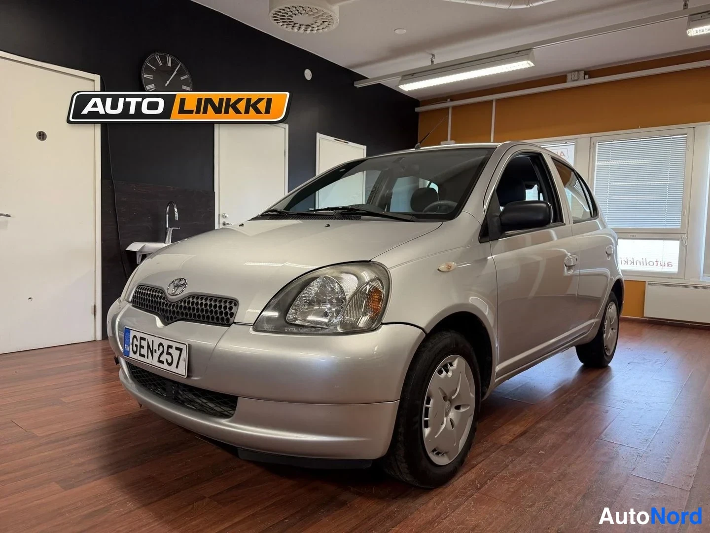 Etuviisto, nokka vasemmalle: Hopea Toyota Yaris -henkilöauto etukulmasta, keula vasemmalla, sisätiloissa.