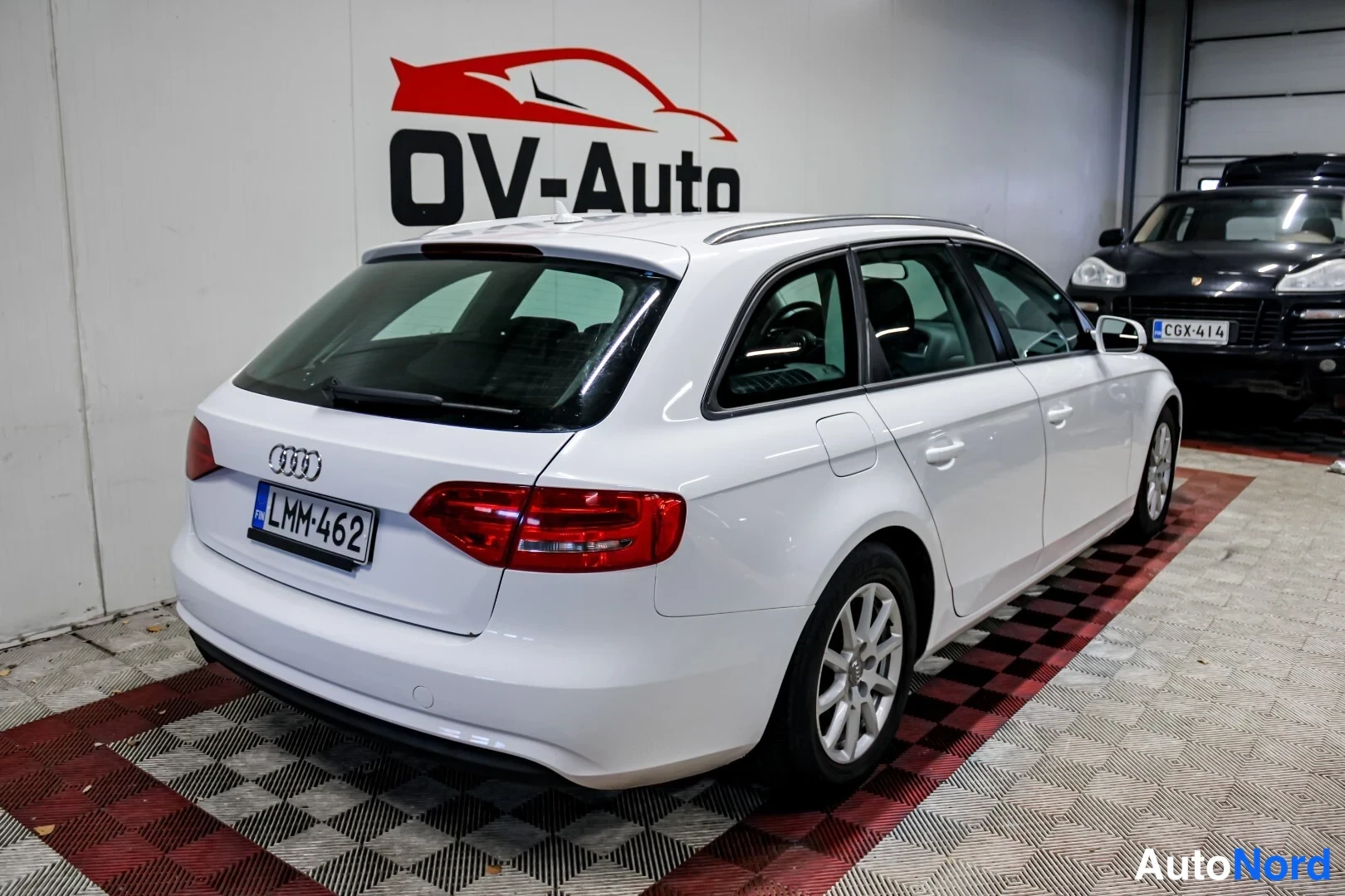 Takaviisto: Auton takapää ja vasen kylki kolmasosan kulmasta: valkoinen farmarimallinen henkilöauto, takaluukussa Audi-merkki, punaiset takavalot ja rekisterikilpi. Kattoon