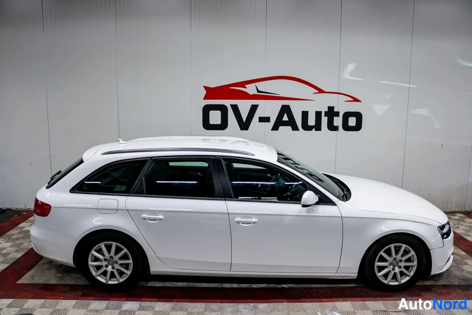 Etuviisto, nokka oikealle: Auton oikea kylki kolmasosan kulmasta nenä oikealla: valkoinen farmarimallinen henkilöauto neljällä ovella, kattokaiteet ja hopeiset kevytmetallivanteet.