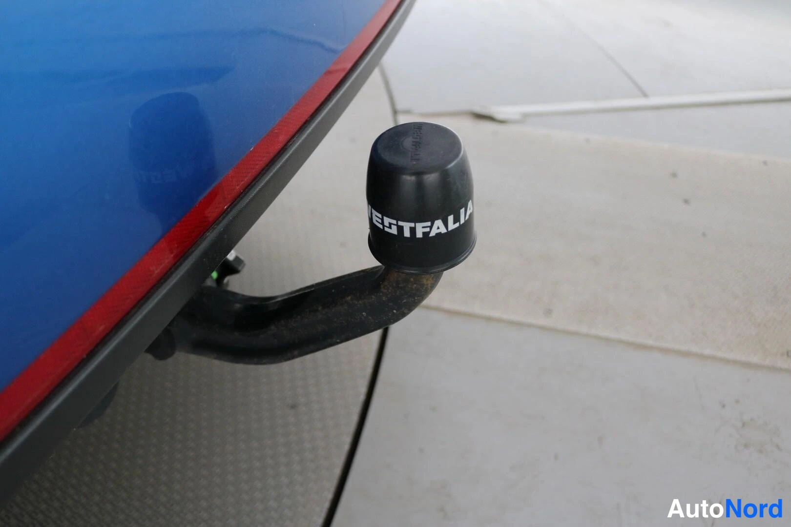 Takaa: Close-up of the car's tow bar with a black protective plug marked 'WESTFALIA'. The car is dark blue with a red stripe on the lower edge of the bumper.