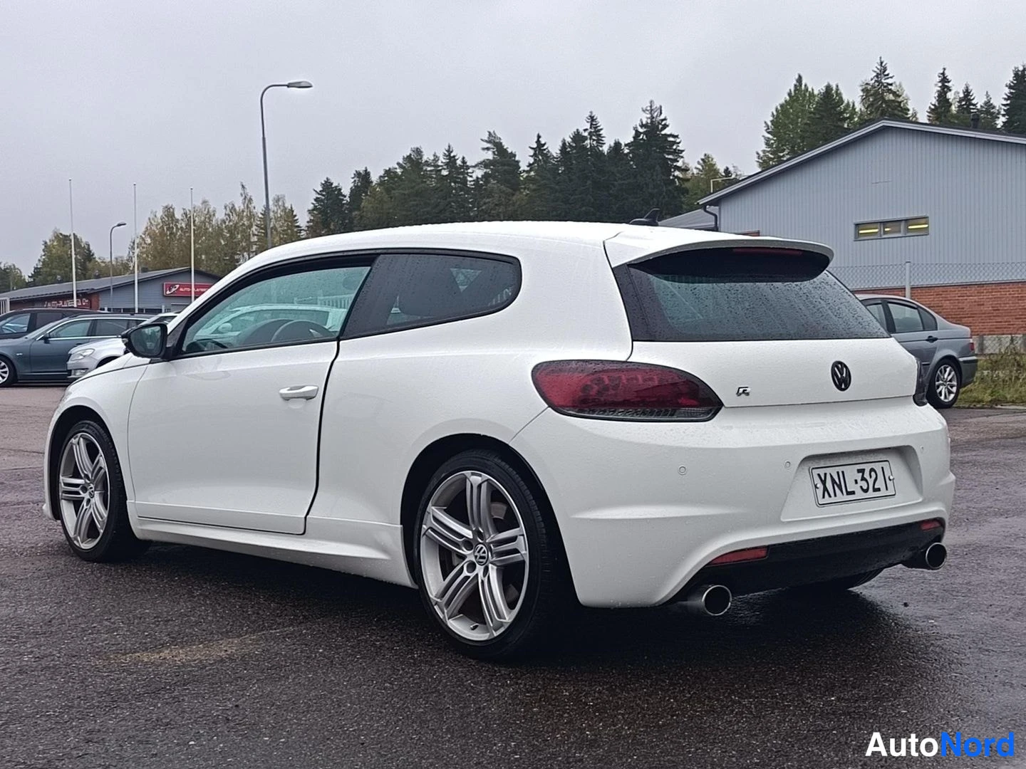 Takaviisto: Valkoinen kolmiovinen Volkswagen-hatchback takaa viistosti kuvattuna; rekisterikilpi, kromivanteet ja kaksiosainen pakoputki näkyvät märällä parkkipaikalla.