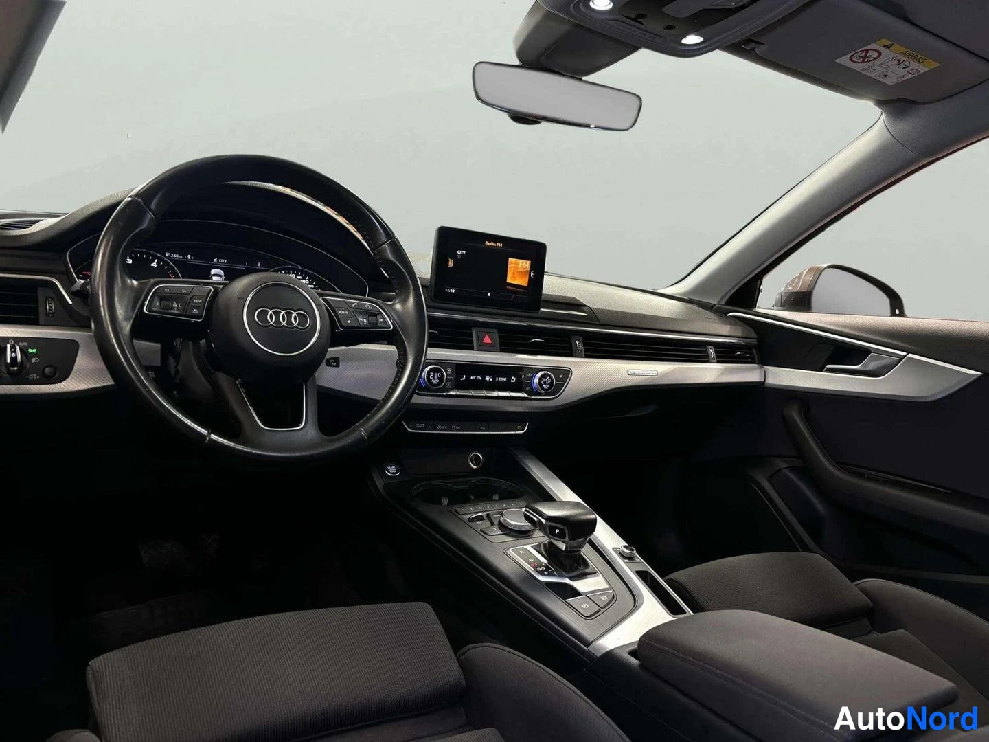 Etuistuimet: Auton sisätilat kuljettajan näkökulmasta: Audi-ohjauspyörä, mittaristo, keskikonsolin automaattivaihteiston valitsin ja käsijarru, tummat kangasistuimet.