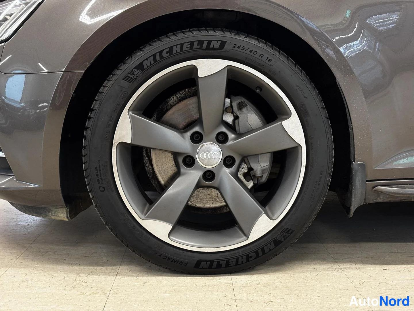 Sivulta: Lähikuva auton etupyörästä, jossa viisimutteroinen Audi-vanteen keskiosa, Michelin-renkuksen kylkiteksti 'MICHELIN' ja merkintä '245/40 R 18' sekä jarrulevy ja