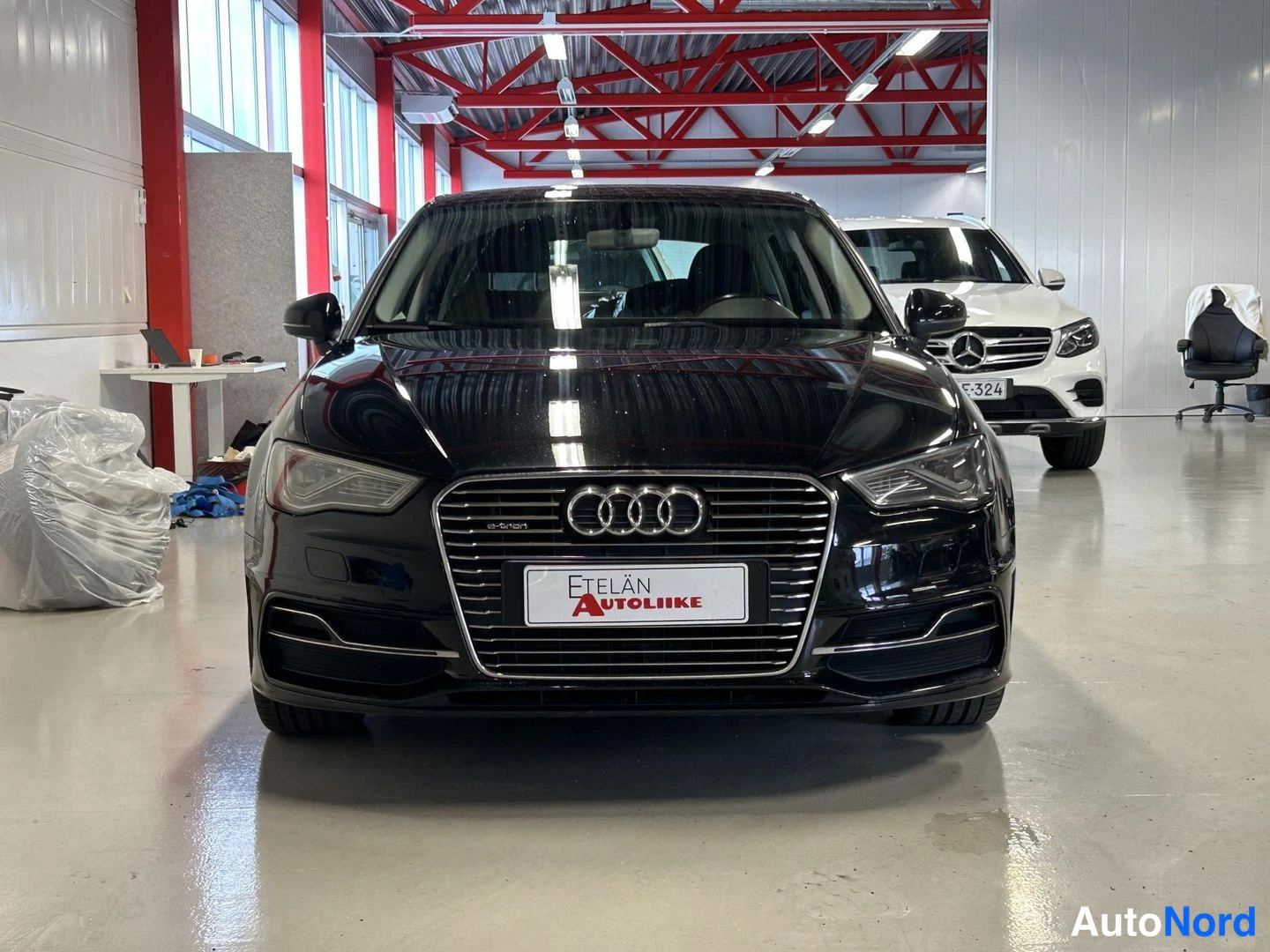 Edestä: Musta Audi A3 suoraan edestä kuvattuna, etusäleikkö ja valojusteet näkyvissä huoltamorakennuksessa, valkoinen Mercedes-Benz taustalla.
