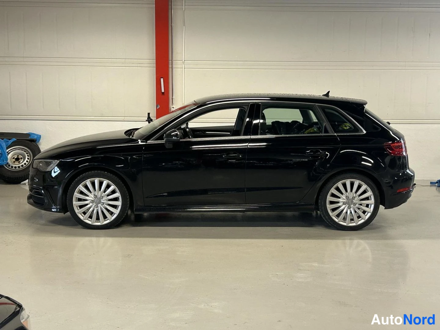 Sivulta: Mustan Audi A3 Sportbackin sivuprofiili vasemmalta, hopeiset vanteet, takaistuimilla näkyvä tavaraa, autotallin sisällä.
