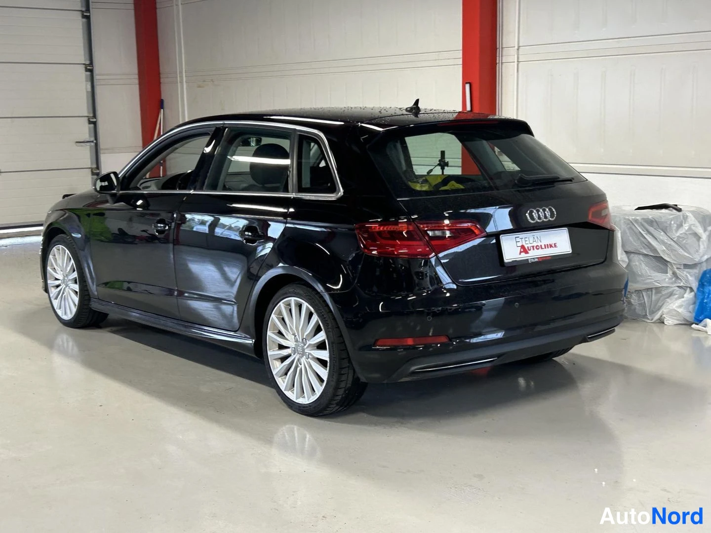 Takaviisto: Mustan Audi A3 Sportbackin takakulmanäkymä sisätiloissa, takavalot ja Audi-merkki näkyvissä, hopeanväriset monihaaravanteet.