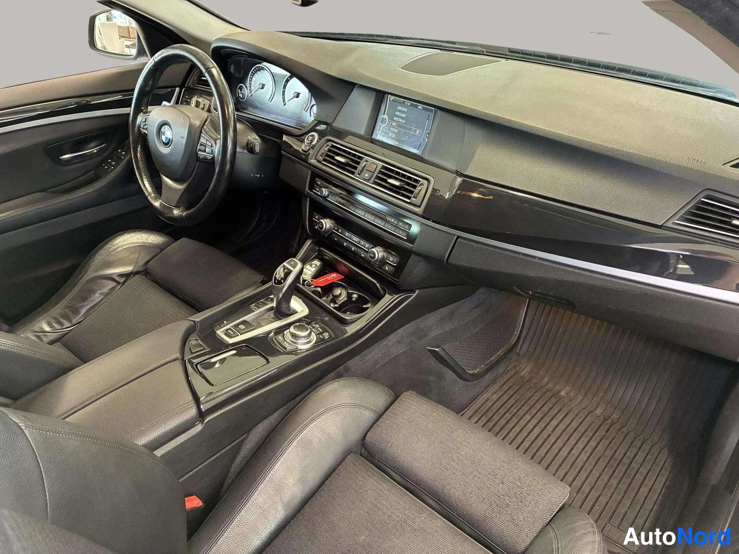 Edestä: Kojelaudan ja keskikonsolin näkymä: BMW-ohjauspyörä, keskellä näyttö, automaattivaihteen valitsin ja iDrive-ohjain. Matot lattiassa.