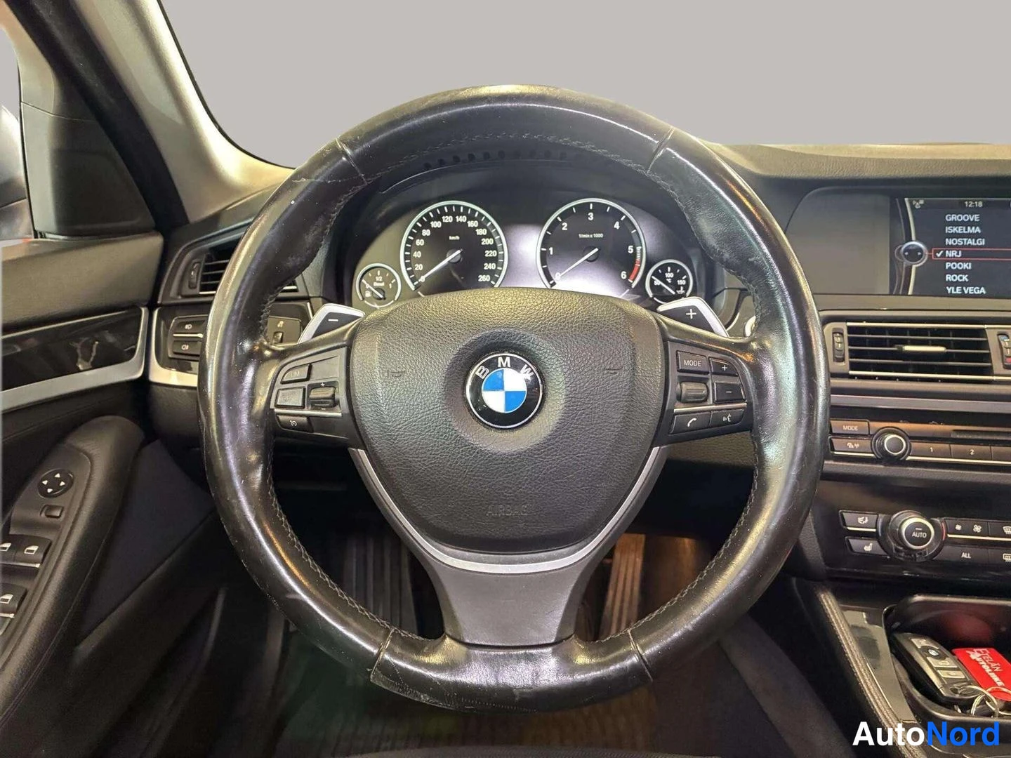 Muu: BMW-merkkisen ohjauspyörän ja kojelaudan näkymä; ratin takana vaihtoahdimet ja näytössä radiokanavalista.