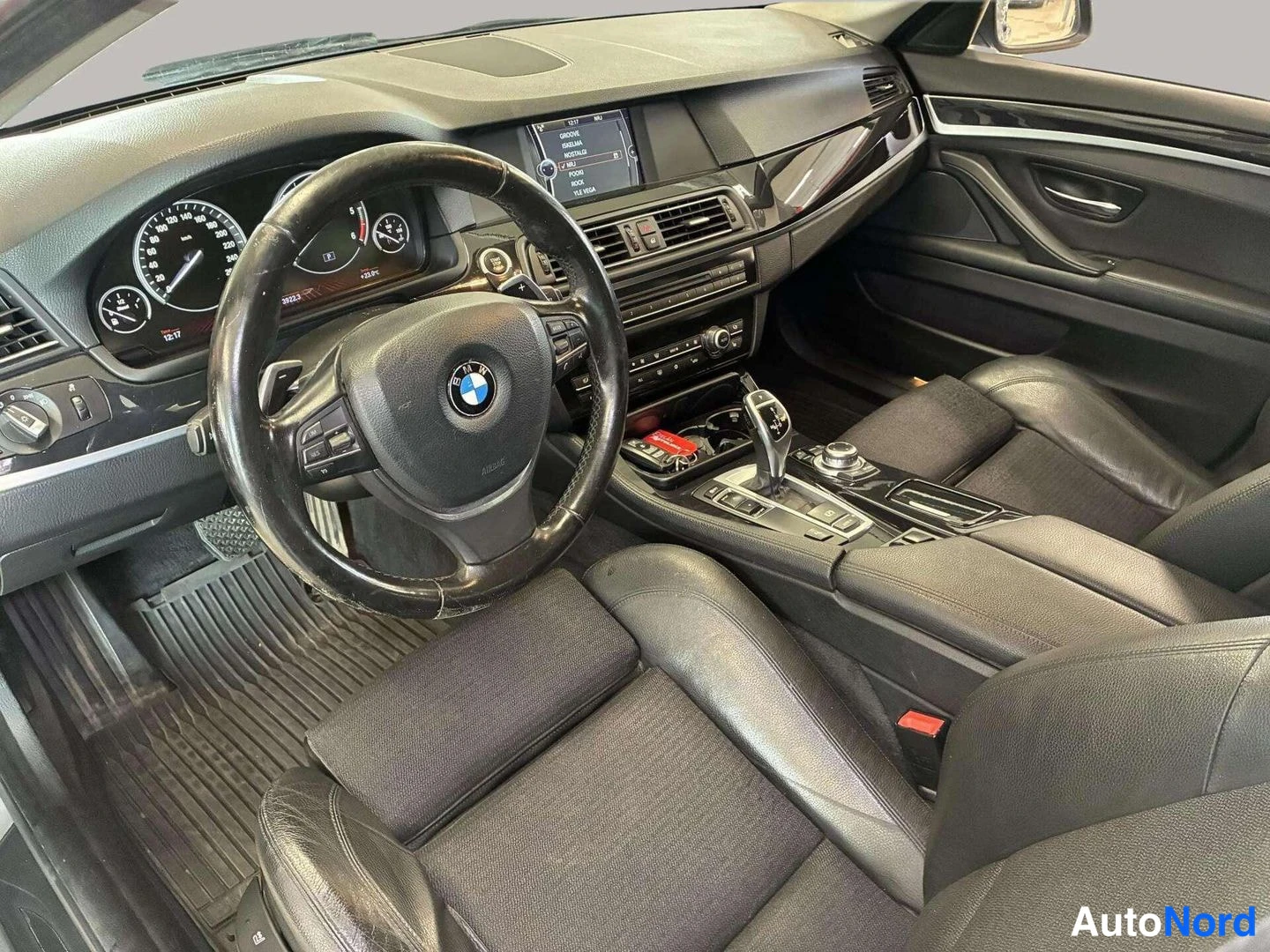 Edestä: Kuljettajan puolen näkymä: BMW-merkkinen ohjauspyörä, mittaristo, keskellä infotainment-näyttö, automaattivaihteen valitsin ja iDrive-ohjain keskikonsolissa.
