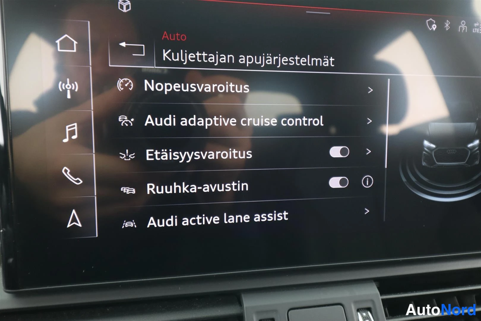 Ohjaamo: Kuvassa on näyttö ruutu, jossa lukee 'Kuljettajan apujärjestelmät'. Näytöllä on listattuna 'Audi adaptive cruise control' ja 'Audi active lane assist' asetusval
