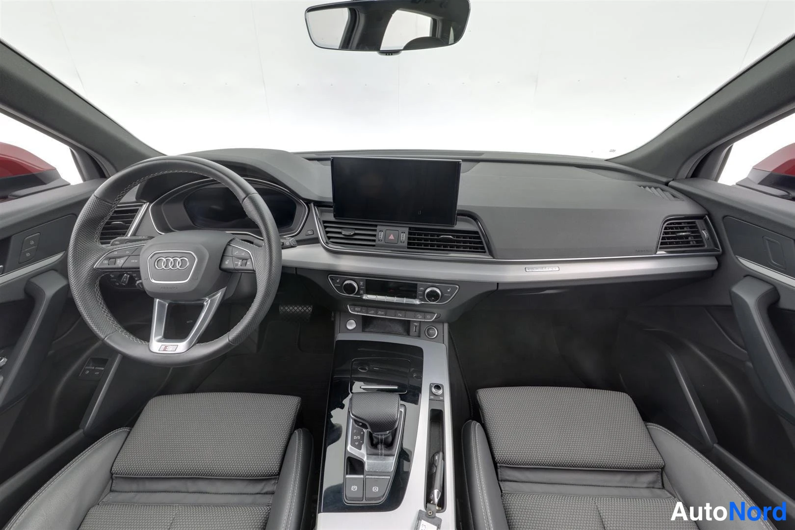 Edestä: Etukenen kojelauta, Audi-merkkinen ohjauspyörä ja korista keskikonsoli. Etupenkkien selkänojat ja musta verhoilu näkyvät.